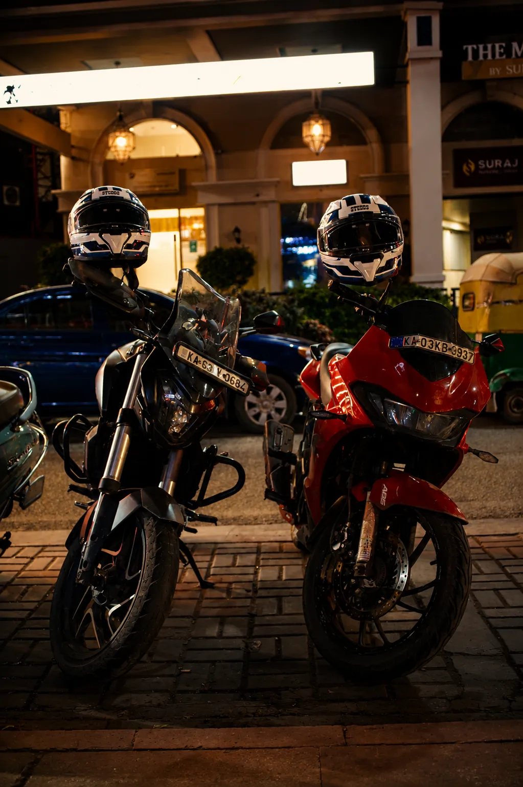 Two motorcycles, one black and one red, parked at night outside a lit storefront with helmets on the seats.