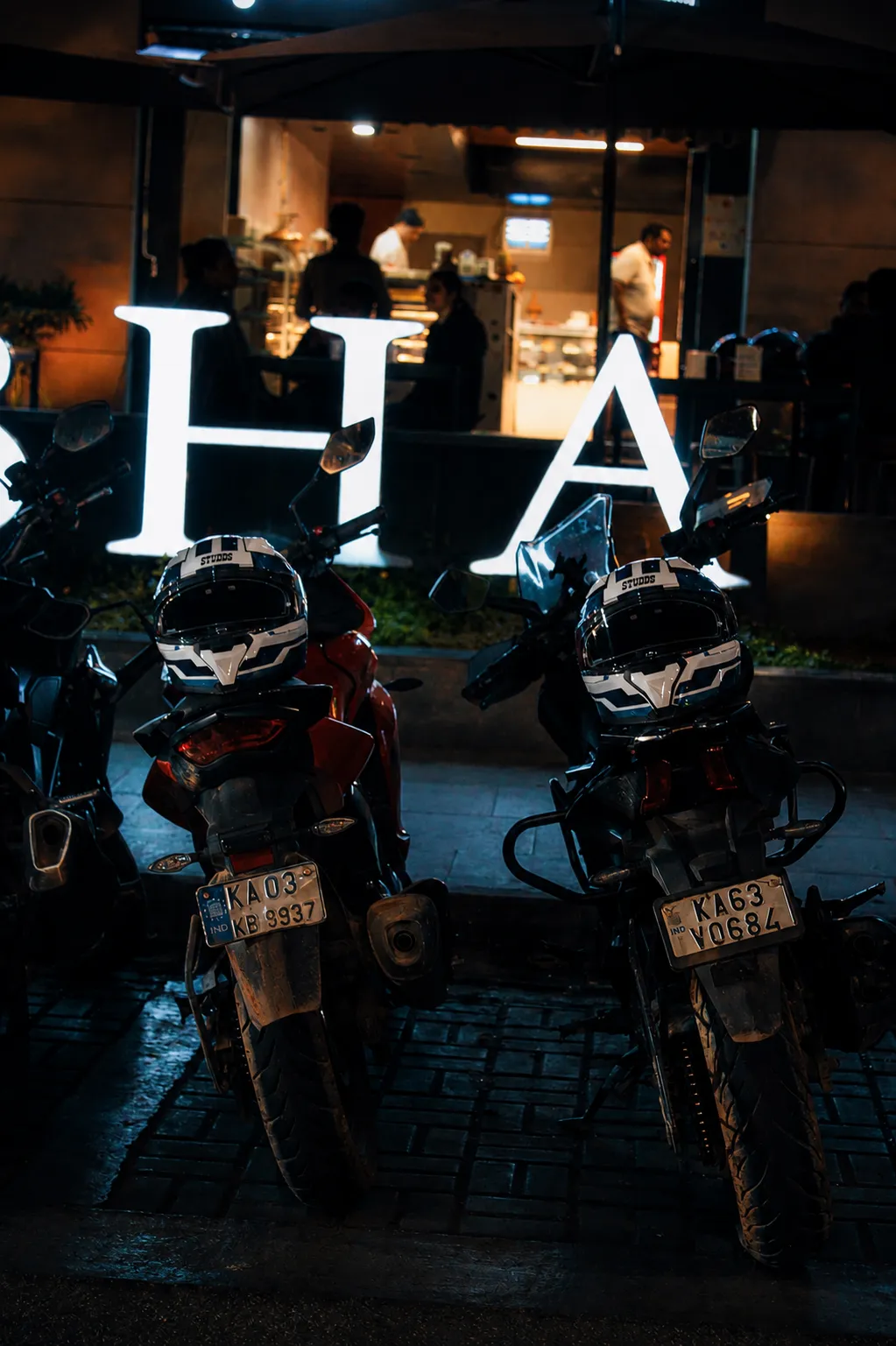 Two motorcycles parked at night in front of a lit sign, helmets on the seats.