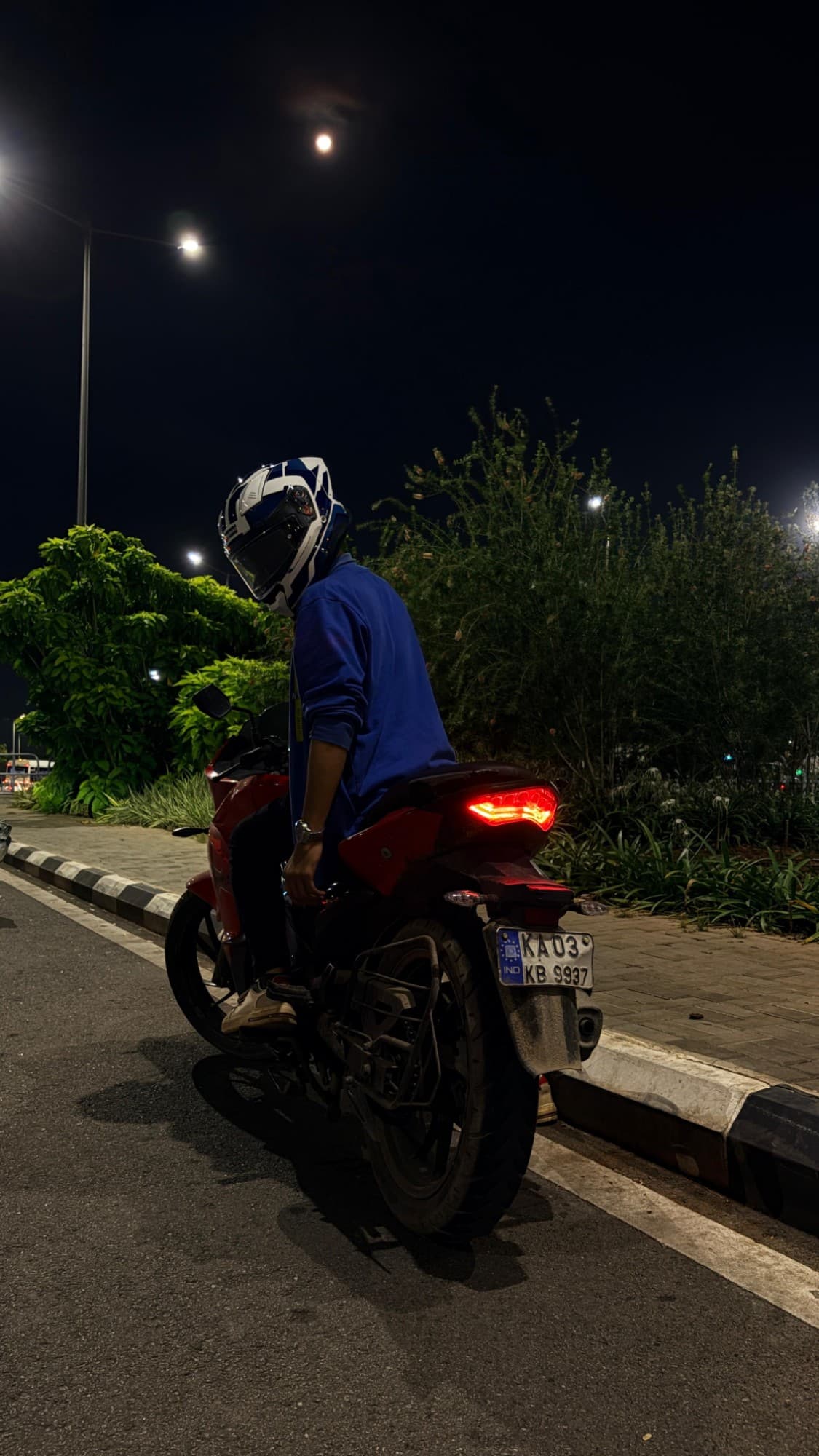 A red motorcycle parked at night on a Bengaluru street, tail light glowing under a streetlamp and a full moon.